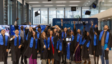 MSc Engineers for Smart Cities graduates in black caps and gowns with blue stoles celebrate their graduation at Université Côte d'Azur, joyfully throwing their caps in the air. One graduate participates remotely via a telepresence robot, visible in the center of the group.