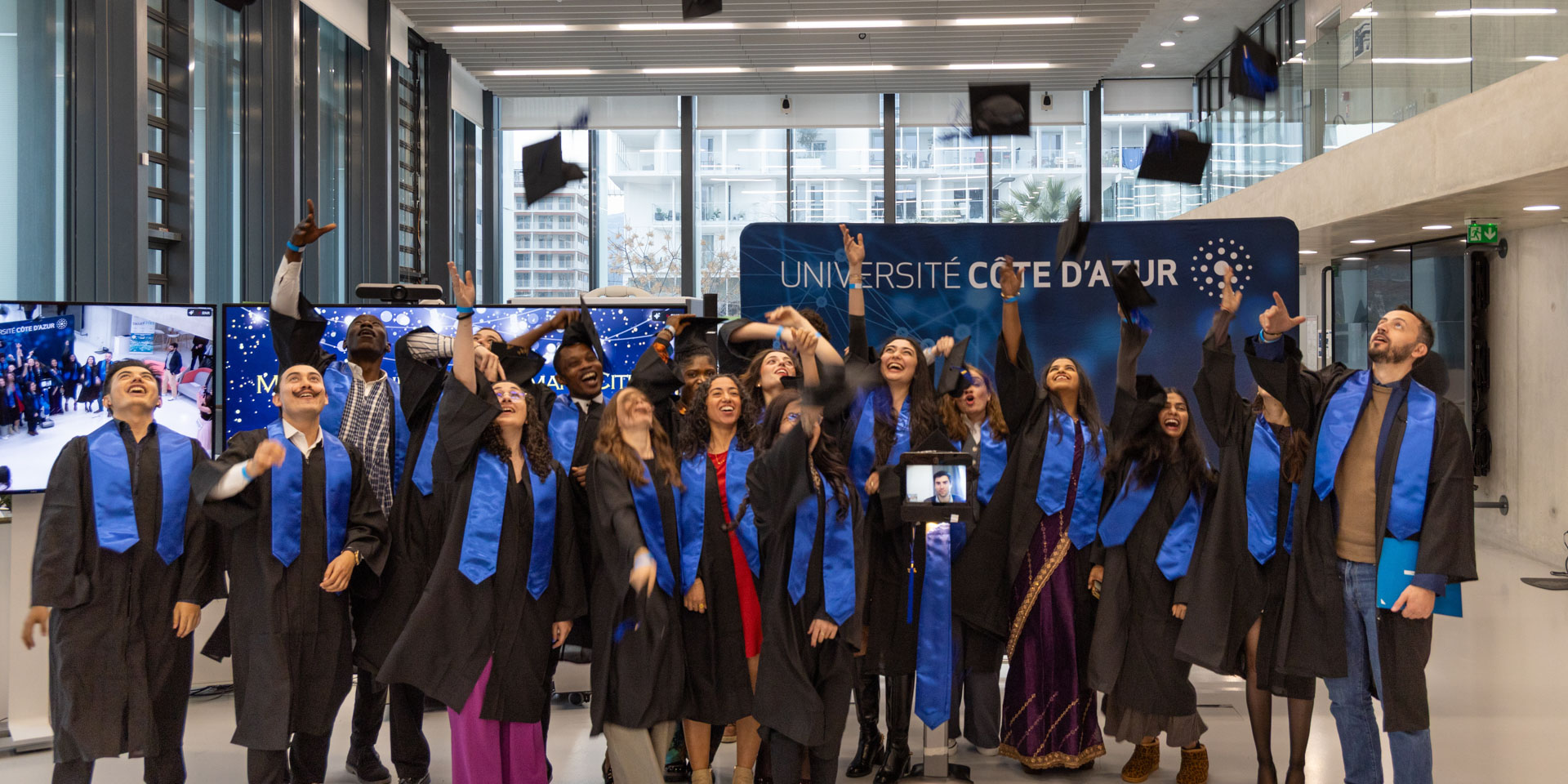 MSc Engineers for Smart Cities graduates in black caps and gowns with blue stoles celebrate their graduation at Université Côte d'Azur, joyfully throwing their caps in the air. One graduate participates remotely via a telepresence robot, visible in the center of the group.